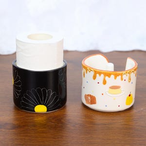 Puede incluir: Dos recipientes de cerámica. Uno es negro con diseños de margaritas blancas y contiene un rollo de papel higiénico blanco. El otro es blanco con un glaseado naranja goteando e ilustraciones de comida.