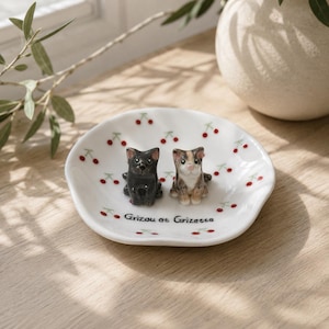 May include: A white ceramic trinket dish with a scalloped edge, decorated with a red cherry pattern. Two small cat figurines, one black and one calico, sit in the center. The text Grizou et Grizette is printed in black on the surface of the dish.