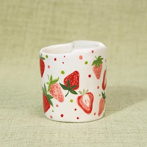 May include: A white ceramic creamer with a cut-out handle, decorated with hand-painted strawberries and green leaves. Red, pink, and green polka dots are scattered around the strawberries. The creamer is a charming addition to any kitchen.
