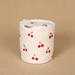 Puede incluir: Un portacepillos de cerámica blanca con un patrón repetido de cerezas rojas y tallos verdes. El soporte tiene una parte superior curvada con una ranura para un cepillo de dientes. El diseño es sencillo y encantador.