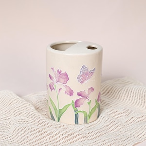 May include: A beige ceramic toothbrush holder with a floral and butterfly design. The holder features purple irises and a butterfly, with green leaves and stems. The holder has a curved top with a small hole for a toothbrush.