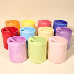 Puede incluir: Una colección de recipientes de cerámica coloridos en varios tonos, incluyendo rojo, naranja, amarillo, verde, azul, rosa y morado. Cada recipiente cilíndrico tiene una abertura curva en el lateral. Los recipientes están dispuestos sobre una superficie clara.