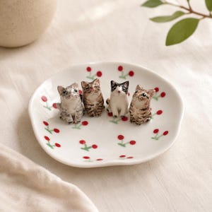 May include: A white ceramic dish with a red cherry pattern featuring four small, hand-painted cat figurines. The cats include a grey tabby, a brown tabby, a black and white cat, and another brown tabby, all sitting upright on the dish surface.