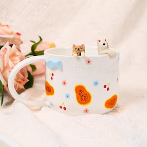 Puede incluir: Taza de cerámica blanca con asa, decorada con rodajas de papaya coloridas, flores y cerezas. La taza presenta una pequeña figura de perro y gato en el borde, y el texto "BOBBY & MAX". También hay un acento azul en forma de pez.