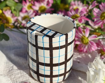 Puede incluir: Un recipiente de cerámica blanco con un patrón de cuadrícula negra y azul. El recipiente tiene una abertura rectangular y una característica similar a un asa. El fondo incluye flores rosas y blancas, creando un contraste vibrante.