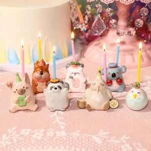 May include: A collection of seven small ceramic animal figurines, each serving as a candle holder with a thin, lit birthday candle. The figures include a pig, a bear, a panda, a koala, and a bird, featuring pastel colors and party hats.