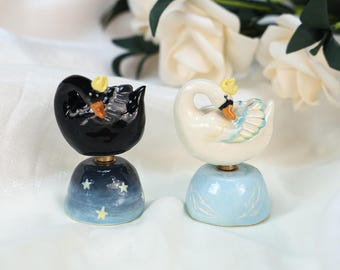Puede incluir: Dos figuras de cisnes de cerámica. Uno es negro, el otro blanco. Ambos tienen coronas amarillas y están sobre bases azules. La base del cisne negro tiene un diseño de noche estrellada. La base del cisne blanco es azul claro.