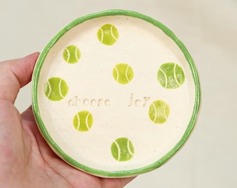 May include: A small, round ceramic dish with a cream-colored interior and a green rim. The dish is decorated with green tennis ball designs and the words "choose joy". The dish is held in a person's hand.