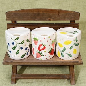 May include: Three white ceramic containers with fruit designs, including olives, strawberries, and lemons. The containers are cylindrical with a small opening at the top and are arranged on a small wooden bench.