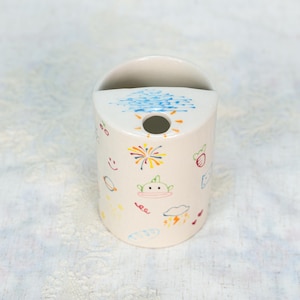 Könnte beinhalten: Ein zylindrischer, cremefarbener Keramik-Zahnbürstenhalter mit gewölbter Oberseite und rundem Loch. Der Halter ist mit farbenfrohen, skurrilen Zeichnungen von Sonne, Wolken, Feuerwerk und einem lächelnden Alien verziert. Das Design ist handgezeichnet.