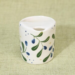 May include: White ceramic toothbrush holder with a curved top and a small hole. The holder is decorated with green olive branches and blue olives. Small blue dots and gold crosses are scattered around the design.