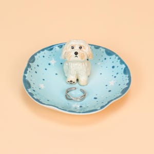 May include: A ceramic trinket dish with a light blue background and white star accents. A small white dog figurine sits in the centre, with a silver ring resting on the dish. The dish has a wavy edge.