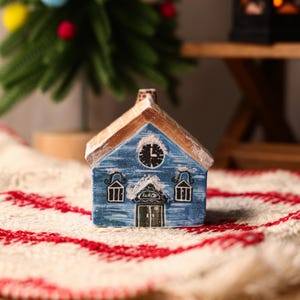 May include: A blue ceramic house with a brown roof and chimney, featuring a clock and two windows. The house is decorated with white accents and sits on a red and white patterned surface. A small Christmas tree is in the background.
