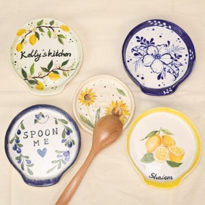 May include: A collection of ceramic spoon rests with various designs. One features lemons and the word "Shalom", another has sunflowers, and a third has the text "Spoon Me". Other rests display floral and lemon branch motifs.