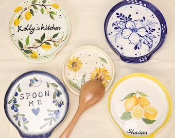 May include: A collection of ceramic spoon rests with various designs. One features lemons and the word "Shalom", another has sunflowers, and a third has the text "Spoon Me". Other rests display floral and lemon branch motifs.