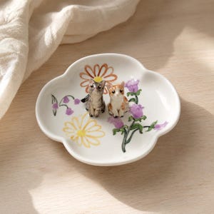 Può includere: Un piattino in ceramica bianca a forma di quadrifoglio, decorato con margherite dipinte a mano in giallo e arancione e fiori viola. Due piccole statuine di gatto dettagliate, una grigia tigrata e una arancione e bianca, si trovano al centro.