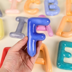 Puede incluir: Una mano sosteniendo la letra "F" azul de un conjunto de letras del alfabeto de cerámica coloridas. Otras letras en el fondo incluyen naranja, rosa y turquesa. Las letras tienen un acabado brillante y un diseño redondeado y juguetón.