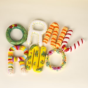 May include: A collection of ceramic letters and shapes spelling "RAW LOVE". The letters are adorned with floral patterns, stripes, and other designs. The letters are in various colours, including green, yellow, orange, red, and white.