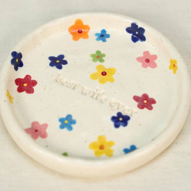 May include: A small, round, off-white ceramic dish decorated with colorful hand-painted flowers. The dish has a raised edge and the words "best wife ever" are written in the center. The flowers are in shades of red, blue, yellow, orange, and pink.