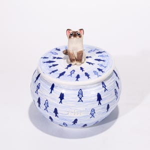 May include: A ceramic jar with a lid, featuring a Siamese cat figurine on top. The jar is white with blue fish illustrations and a blue and white striped pattern. The cat figurine is brown and tan.