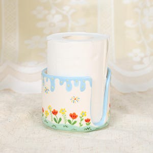 Puede incluir: Un portarrollos de papel higiénico de cerámica con un diseño floral. El soporte es blanco con un borde azul y una base verde. Flores rojas, amarillas y naranjas están pintadas en el soporte. Un rollo de papel higiénico blanco está dentro.