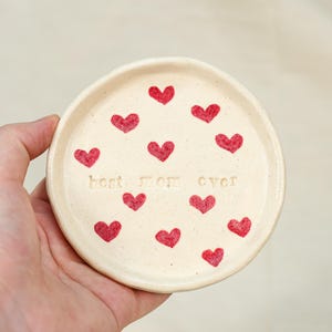 May include: A small, off-white ceramic dish with a slightly raised edge. The dish is decorated with red heart shapes and the words "best mom ever" in the centre. The dish is held in a hand.