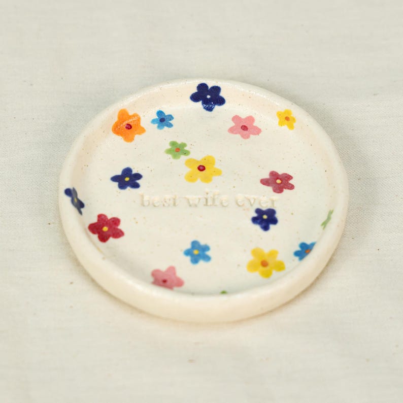 May include: A small, off-white ceramic dish decorated with colorful flowers in shades of orange, blue, pink, yellow, and green. The words "Best Wife Ever" are inscribed in the center of the dish.