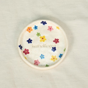 May include: A small, off-white ceramic dish decorated with colorful painted flowers in shades of orange, pink, blue, yellow, and green. The words "best wife ever" are inscribed in the center. The dish has a slightly raised edge.