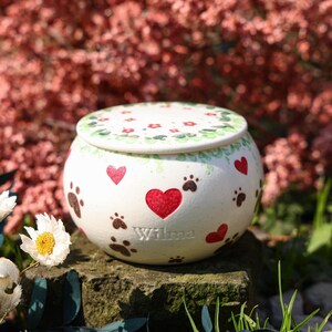 Può includere: Urna per animali domestici in ceramica bianca con coperchio, decorata con cuori rossi e impronte di zampe marroni. Il nome "Wilma" è inciso sull'urna. Il coperchio presenta un motivo floreale con fiori rossi e foglie verdi.