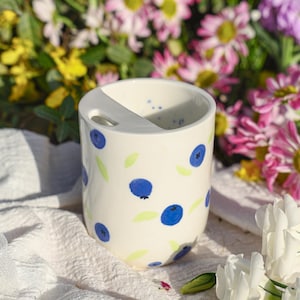 May include: A white ceramic utensil holder with a blueberry pattern. The holder has a divided interior and a handle. Blue blueberries and green leaves are painted on the exterior. The background features colorful flowers.