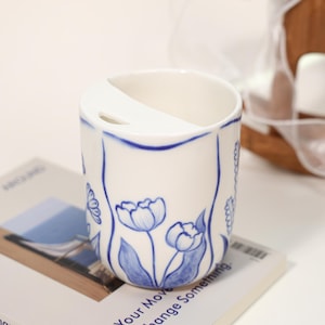 May include: A white ceramic toothbrush holder with blue floral designs. The holder has a curved divider and a blue trim. The design includes blue tulip illustrations. The holder is on a book with the text "Your Move Change Something."
