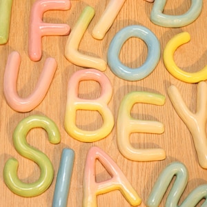 Könnte beinhalten: Eine Sammlung bunter Keramikbuchstaben in verschiedenen Pastelltönen, darunter Rosa, Gelb, Grün und Blau. Die Buchstaben sind auf einer hellbraunen Holzoberfläche angeordnet und bilden eine verspielte und dekorative Darstellung. Die Buchstaben sind in einer Vielzahl von Schriftarten.