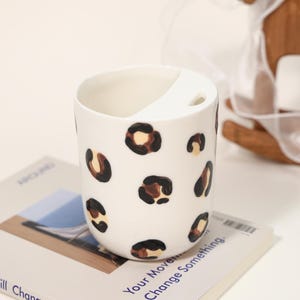 Puede incluir: Taza de cerámica blanca con un diseño de asa único, adornada con un estampado de leopardo en marrón y negro. La taza está sobre un libro con el texto "Your Move. Change Something."