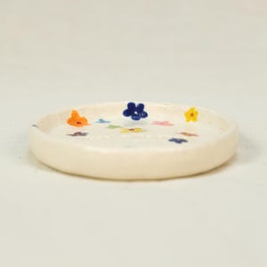 May include: A small, off-white ceramic dish with a slightly raised rim. The dish is decorated with colorful, hand-painted flowers in shades of orange, blue, yellow, pink, and green. The dish is round and appears to be handmade.