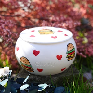 May include: A white ceramic jar with a lid, decorated with red hearts and cartoon burger illustrations. The jar has the name "Charly" inscribed on the side. The jar is sitting on a stone.