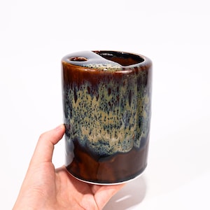 May include: A cylindrical ceramic toothbrush holder with a dark brown, blue, and tan glaze. The top has a small hole and a larger opening. The holder is held by a hand against a white background.