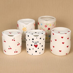 May include: Five white ceramic containers with various designs. One has cherries, another has polka dots and the text "Be with ME" with a heart, and others have floral patterns and hearts. Each container has a small opening on top.