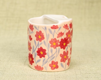 May include: A ceramic toothbrush holder with a light peach background and a floral pattern of red flowers with yellow centers and blue stems. The holder has a handle and a compartment for toothbrushes.