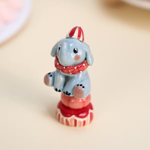 Könnte beinhalten: Hellblaue Keramik-Elefantenfigur mit rot-weiß gestreiftem Partyhut und rotem Kragen. Der Elefant sitzt auf einem rot-rosa dekorativen Sockel. Die Figur hat schwarze Flecken auf den Füßen.