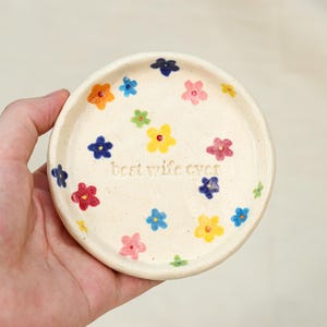 May include: A small, round, off-white ceramic dish decorated with colorful hand-painted flowers. The dish has the phrase "best wife ever" inscribed in the center. The flowers are in shades of red, orange, yellow, blue, and pink.