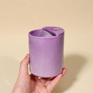 Peut inclure: Un vase cylindrique en céramique lavande avec une découpe en forme de cœur sur le bord. Le vase a une finition lisse et brillante et est tenu dans une main. Le design est simple et moderne, adapté aux petits arrangements floraux ou comme pièce décorative.
