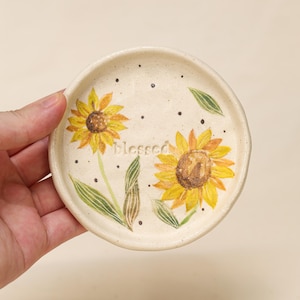 Op de afbeelding: Een kleine, ronde keramische schaal met een crèmekleurige basis. Twee gele zonnebloemen met bruine centra zijn op de schaal geschilderd, samen met groene bladeren en het woord "blessed". Zwarte stippen zijn verspreid rond de zonnebloemen.
