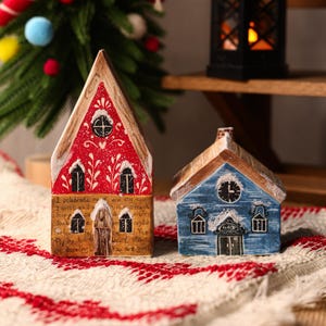 May include: Two decorative miniature houses. One is red with white accents and a text, the other is blue with a clock design. Both have a snow-like effect on the roof and are placed on a red and white patterned surface.