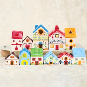 Puede incluir: Una colección de casas en miniatura de cerámica coloridas. Cada casa presenta diseños únicos, incluyendo tejados rojos, azules, verdes y amarillos, con ventanas y puertas detalladas. Las casas están dispuestas sobre una superficie blanca.