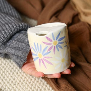 Puede incluir: Un pequeño recipiente de cerámica color crema con una parte superior curvada y una abertura circular. El recipiente está decorado con patrones florales de margaritas azules, moradas y blancas. El recipiente está en la mano de una persona.
