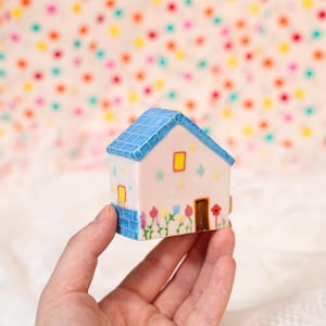 Può includere: Una casa in ceramica in miniatura con tetto blu a tegole, pareti rosa chiaro e una porta marrone. La casa presenta piccole finestre gialle ed è decorata con fiori dipinti. La casa è tenuta in mano.