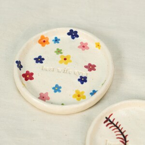 May include: A small, round, off-white ceramic dish decorated with colorful hand-painted flowers. The dish has the text "best wife ever" in the center. The flowers are in shades of blue, pink, yellow, and orange.
