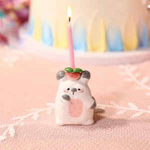 May include: A small, white, bear-shaped candle holder with grey ears and paws, a pink belly patch, and a red strawberry on its head. A thin, lit pink birthday candle is inserted into the top of the strawberry, casting a warm glow.