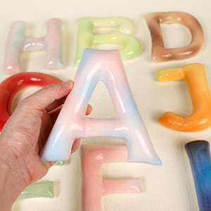 May include: A hand holding a pastel pink and blue letter "A", with other colourful letters in the background. The letters, including "H", "B", "D", "J", and "F", appear to be made of a translucent material. The letters are in various colours.