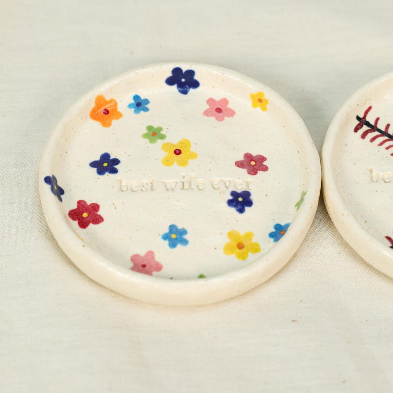 May include: A pair of small, round, off-white ceramic dishes. One dish is decorated with colorful flowers and the text "best wife ever". The flowers are in shades of blue, yellow, orange, and pink.